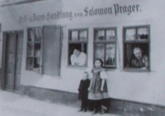 Unscharfes Schwarz-Weiß-Foto des Geschäfts. Zwei Personen schauen aus den Fenstern, davor ein junges Mädchen mit einem Kind an der Hand