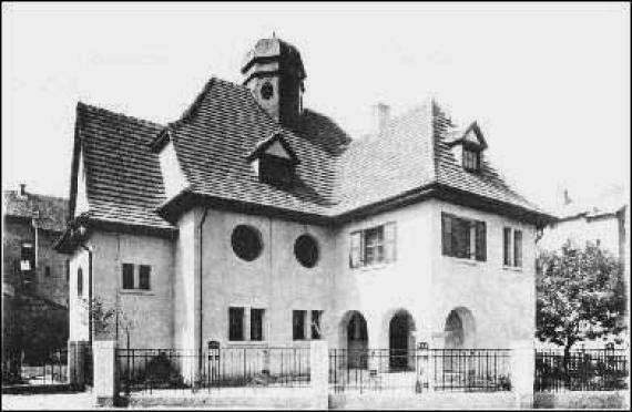 Black and white photo of the synagogue. Striking are two round windows and the entrance, which is reached through three arches.