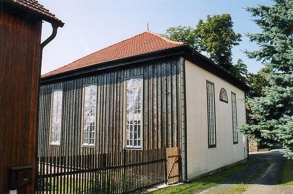 eine Wand vertäfelt, die andere verputzt. Ein einfacher Holzzaun grenzt das Grundstück ein.