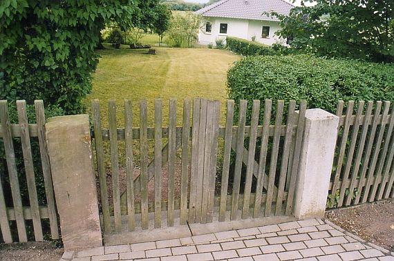 Entrance gate made of wooden slats, behind it lawn, on the left single gravestones, on the right a residential house