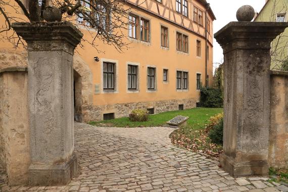 Cobblestones lead to the memorial plaque, which is on the ground. Behind it green lawn, to the left a plastered half-timbered house.