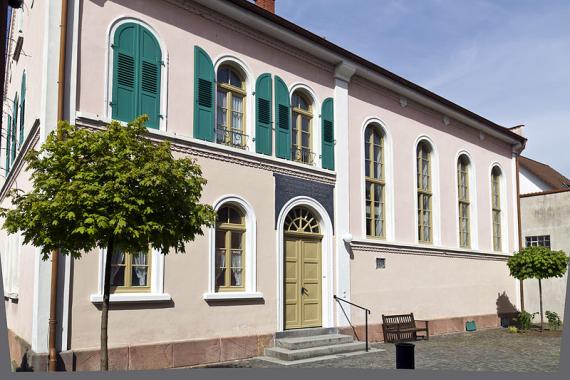 Old pink building with round arch windows, in front of it a small square with cobblestone pavement