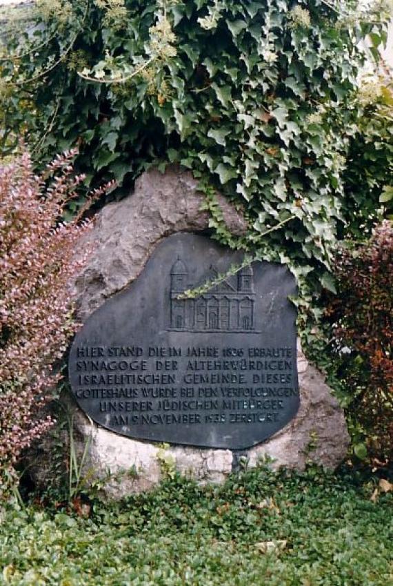 Gendenktafel aus Metall auf einem Stein, umrankt von Pflanzen