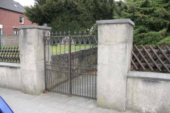 Ansicht des Eingangs in den Friedhof. Niedrige Steinmauer mit Zaun führt ins Grüne. links im Bild ein rotes Haus