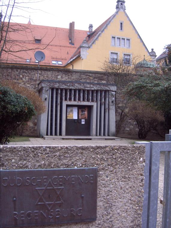 Haus der jüdischen Gemeinde Regensburg in Gelb, vorne Eingangstor aus Metal und auf einer steinernen Wand ein Metallplatte mit der Inschrift: "Jüdische Gemeinde Regensburg"