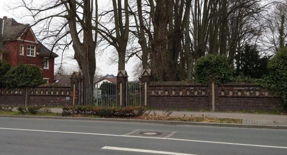 View of the entrance of the cemetery from Massener Straße. The cemetery entrance forms a low wall with red stones. On the left side of the picture a red house and trees