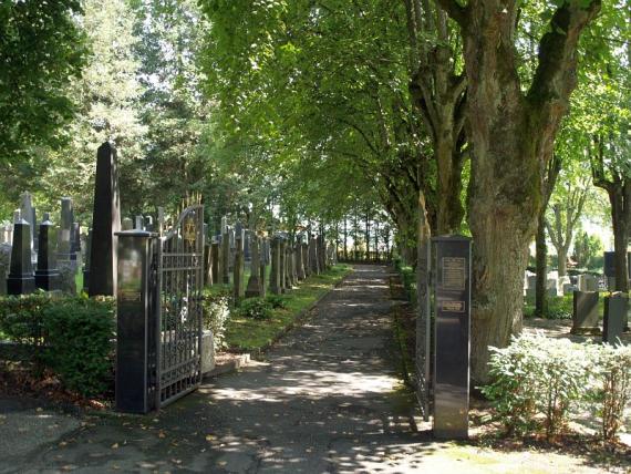 Geöffnetes Tor zu einem Friedhof, viele Bäume und Grabsteine