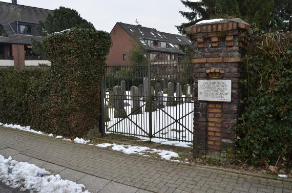 On the right side of the picture a brick column with a memorial plaque, on the left side of the column a fence with a view of the snow covered cemetery