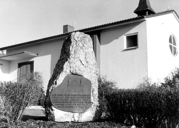 Schwarz-weiß Bild von einem einstöckigen Haus mit abgeschrägtem Dach, davor ein Gedenkstein