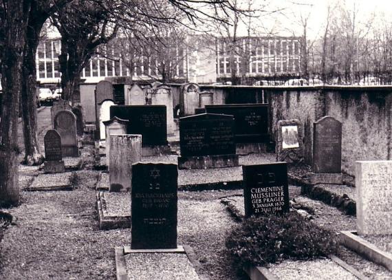 Black and white image of a cemetery, several gravestones, on one side a wall, on the other trees