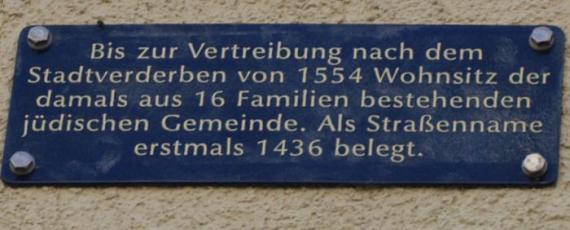 Blaue Hinweistafel mit weißen Lettern: "Bis zur Vertreibung nach dem Stadtverderben von 1554 Wohnsitz der damals aus 16 Familien bestehenden jüdischen Gemeinde. Als Straßenname erstmal 1436 belegt."