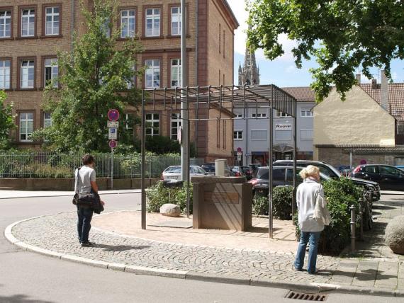 eine Stele unter einem Stahldach in der Mitte eines Platzes. Im Hintergrund links ein großes Gebäude und Bäume