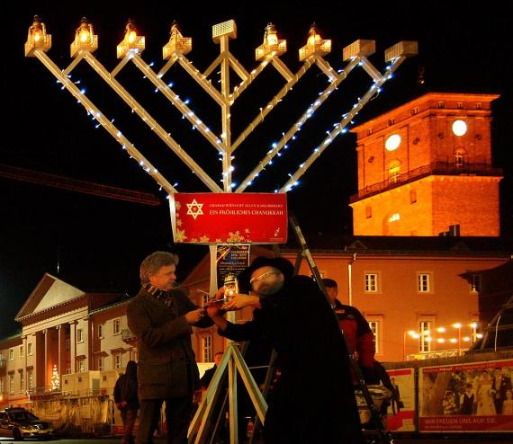 A large multi-armed candelabrum is lit in a public square by two men