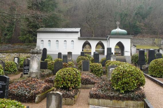 bepflanzten Grabsteine auf dem Friedhof, im Hintergrund ein weißes Haus mit Bögen und einem Davidstern auf einer Kuppel