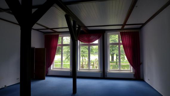 View of interior with blue carpet, three large windows with red curtains.