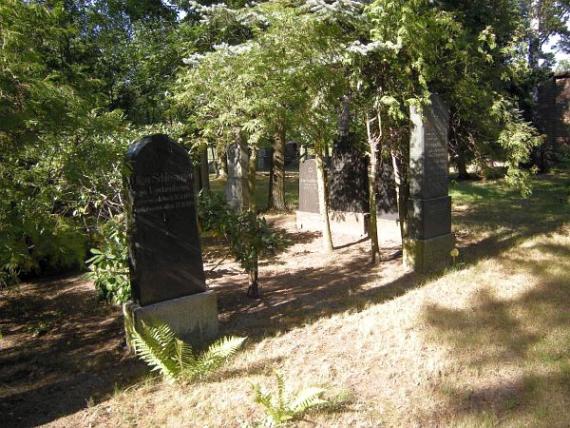 Ein dunkler Grabstein auf dem Friedhof, drum herum Bäume