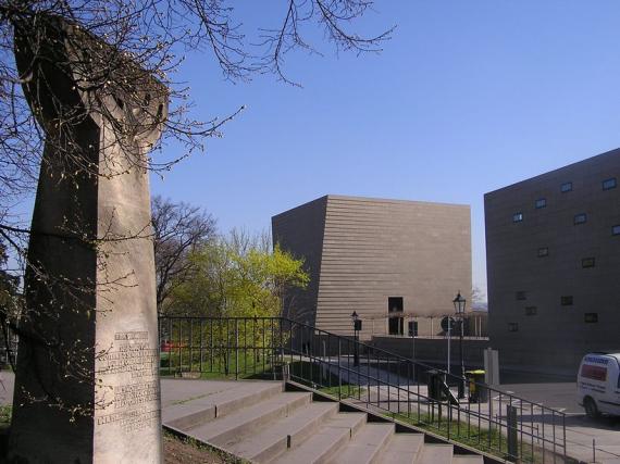 Blick auf den großen, quadratischen Bau der Neuen Synagoge. Links im Bild eine steinerne Stele in Form eines siebenarmigen Leuchters