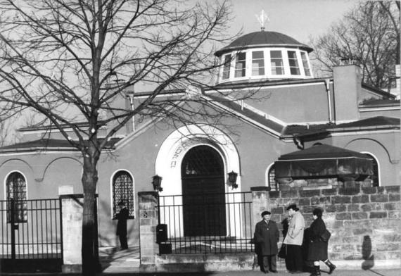 Schwarz-Weiß Bild eines breiten Gebäudes mit einer Kuppel. Eingangstür mit weißen Bogen und hebräischen Lettern. Auf der Straße vor der Synagoge Menschen