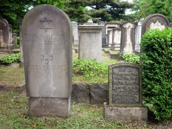Zwei Grabsteine auf dem Friedhof, links ein großer, bogenförmiger Grabstein mit deutsch und hebräischen Inschriften. Rechts ein kleiner, quadratischer Grabstein