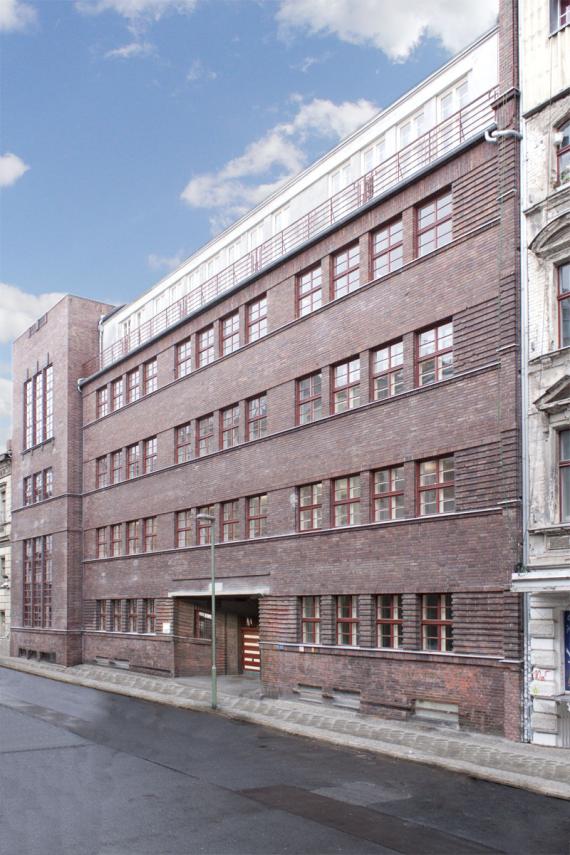Blick von der Straßenseite auf ein großes, mehrstöckiges, langes Gebäude aus rotem Backstein. Das Gebäude wurde als Schule genutzt. Es hat viele Fenster.
