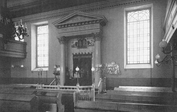Black and white picture of the interior of the synagogue with wooden benches on the right and left and a prayer lectern in the center of the room