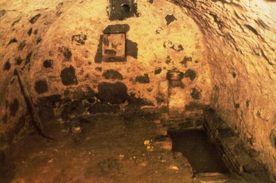 View of the underground mikvah, orange light illuminates the arched, not particularly high, stone walls. A bathtub-sized recess in the floor is in the corner. A niche in the upper center of the picture was probably used to place lamps there.