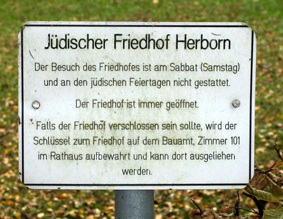 Notice board at the entrance of the cemetery. Text: Herborn Jewish Cemetery. Visiting the cemetery is not allowed on Sabbath (Saturday) and Jewish holidays. The cemetery is always open. If the cemetery is locked, the key to the cemetery is kept at the building office, room 101 in the town hall and can be borrowed there. (text end)