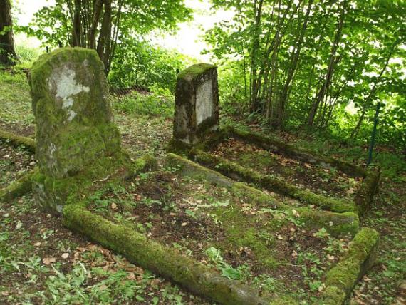 Zwei einzelne moosbewachsene Grabsteine, auf der Erde wächst Unkraut