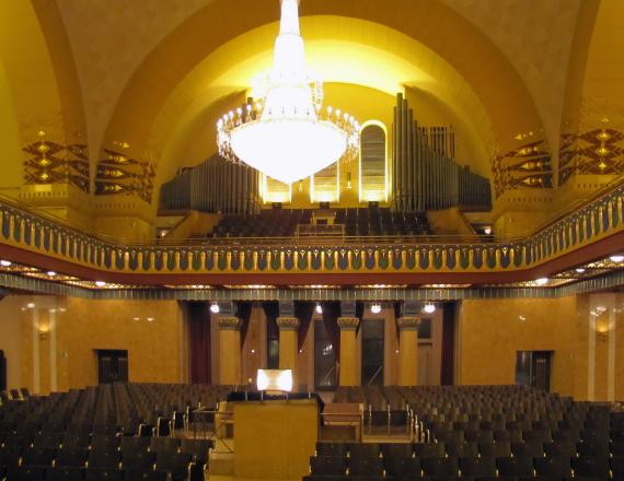 View into the prayer room of the synagogue with chandelier hanging from the ceiling, a large organ in the back. Left and right below wooden benches