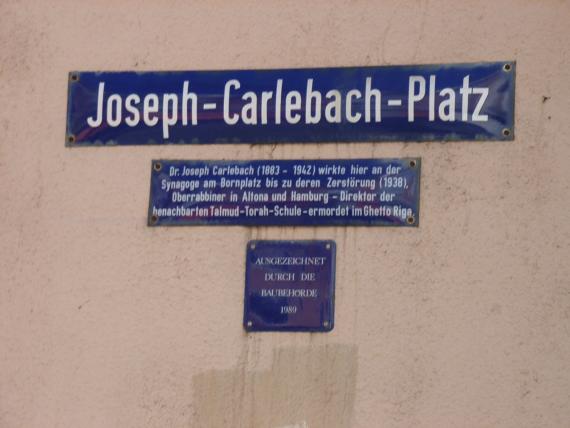 Drei blaue Straßenschilder, das Obere Schild: "Joseph-Carlebach-Platz", darunter ein Schild mit Lebensdaten von Carlebach, darunter ein kleines Schild, das lautet: "ausgezeichnet durch die Baubehörde 1989"