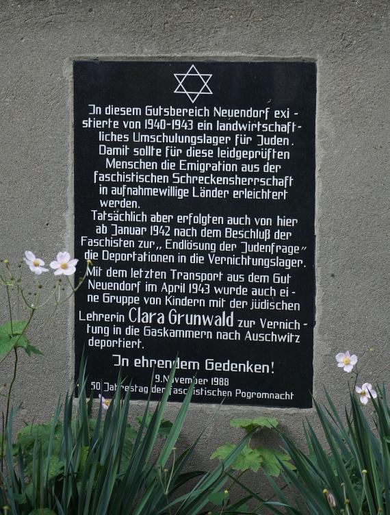 Black plaque with white letters in memory of the Jewish workers' colony on a concrete wall.