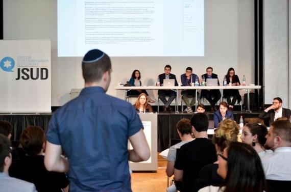 Großer Versammlungsraum mit mehreren Personen, im Vordergrund steht eine Person mit dem Rücken zur Kamera, im Hintergrund sitzen mehrere Personen auf dem Podium.