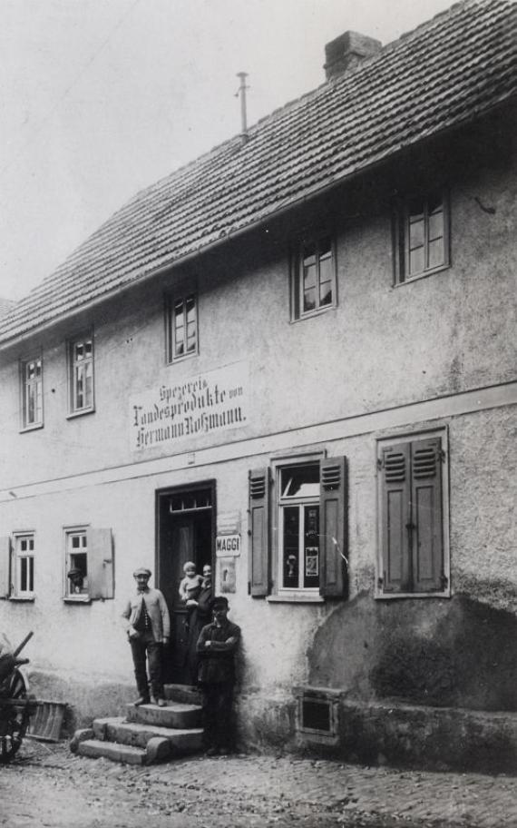 Black and white shot of the Rossmann family home, three people with a baby are standing in the entrance of a simple apartment building, another person is looking out of the window on the first floor. Open shutters decorate the windows. The house is located on the main street.