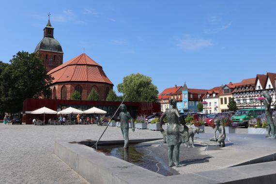 Large market square with a fountain and stone statues, in the background a church