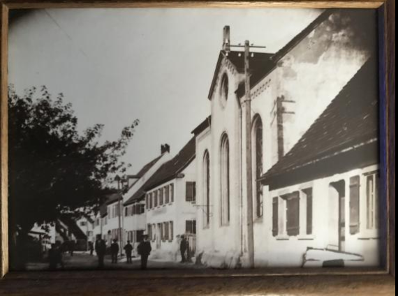 Black and white picture of a synagogue in Schopfloch. The building is on the right side of the picture. In front of it some passers-by are standing on the street. On the left there is a tree.