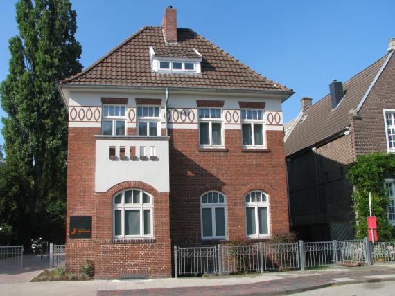 The picture shows a family house built around the turn of the century. It is a red brick house with a brown roof and a small bay window with a balcony on top. Both the upper part of the bay window and the upper part of the upper floor are plastered in white. The upper floor also has pointed vertical ovals as decoration around the building under the roof.