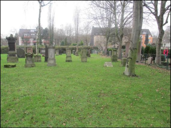 View of a meadow. Some gravestones, scattered. Behind a wall, behind houses.