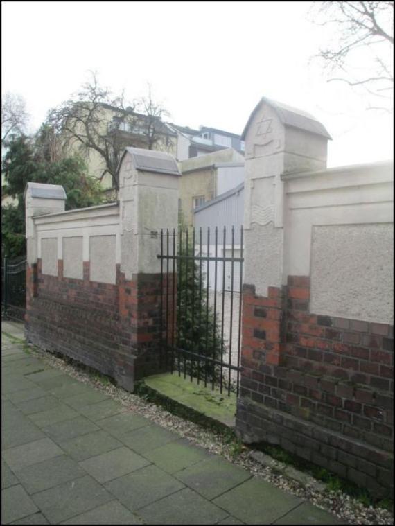 Man sieht eine hohe Backsteinmauer mit einem Gittertor. Oben an einem Pfeiler der Magen David, der Davidstern.