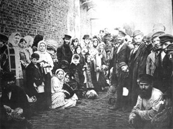 Gruppe von Männern, Frauen und Kindern stehen versammelt in einer schmalen Straße, links eine Ziegelwand, alle blicken in die Kamera.