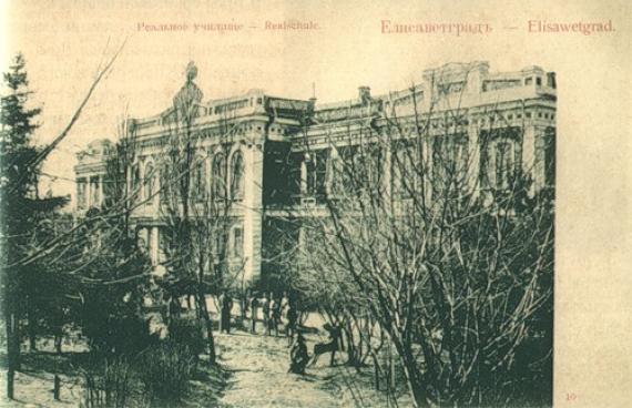 Old picture postcard with the view of a building, on the top Russian and German inscription "Realschule - Elisawetgrad". In front of the building a park with trees partially covering the view of the building.