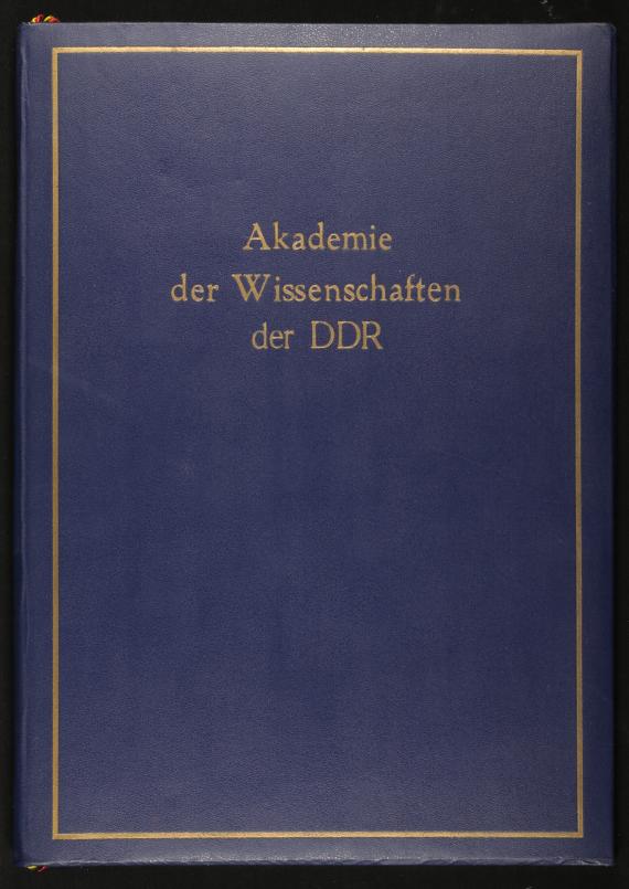 Eine blaue Mappe, die gold mit dem Text “Akademie der Wissenschaften der DDR” bedruckt ist; die Mappe enthält eine Urkunde, auf der mit großen Lettern Lola Zahn für ihre 25jährige Mitgliedschaft ausgezeichnet wird. 