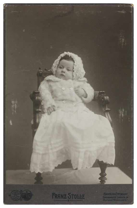 Fotografie in schwarz-weiß, auf welchem Helene als Kleinkind auf einem Stuhl sitzend zu sehen ist. Sie trägt ein weißes Kleid, kombiniert mit einem weißen Jäckchen und einer ebenso weißen Mütze. Ihr Haar ist brünett und ihr Blick freundlich.