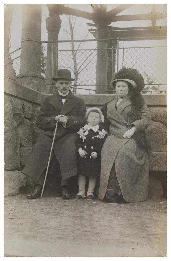 Fotografie in schwarz-weiß, auf welchem Helene als Kleinkind zwischen ihrem Vater (links sitzend) und ihrer Mutter (rechts sitzend) stehend zu sehen ist. Alle drei sind vornehm angezogen, der Vater mit Hut und Gehstock, die Mutter mit Hut und Pelzstola. Beide gucken mit einem neutralen Blick in die Kamera. Helenes Gesicht ist leicht verschwommen.