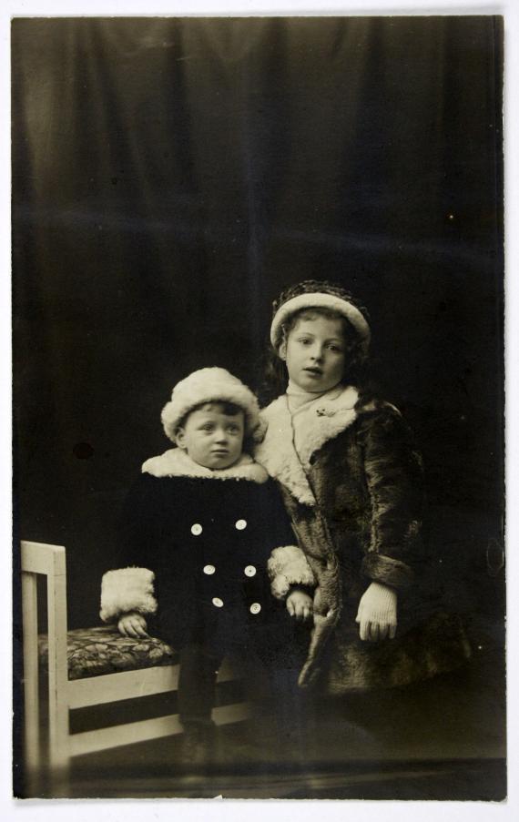 Fotografie in schwarz-weiß, auf welchem die junge Helene gemeinsam mit ihrem jüngeren Bruder Victor auf einer Bank sitzend in einem Foto-Atelier zu sehen sind. Sie sehen beide leicht an der Kamera vorbei und haben Wintermäntel und Mützen auf. 