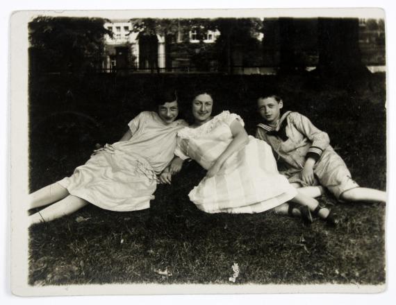 Fotografie in schwarz-weiß, auf welchem Helene (links) mit einem Mädchen und einem Jungen auf einer Wiese liegend zu sehen sind. Die Mädchen tragen beide weiße Kleider, der Junge eine Art Uniform, hell gestreift. Alle drei blicken lächelnd in die Kamera.