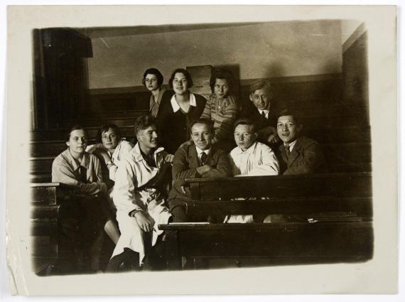 Photography in black and white, on which ten students can be seen in a lecture hall, both sitting and standing. Most of them are smiling into the camera, Helene, who can be seen sitting in the far left background, is looking in another direction with a neutral gaze.