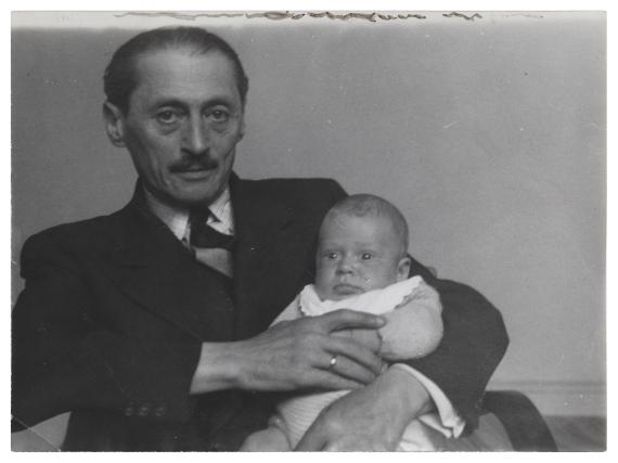 Schwarz-weiß-Fotografie, auf welchem ein älterer Mann in einer sitzenden Position zu sehen ist. Er hält ein pummeliges Baby in den Armen und schaut direkt in die Kamera.