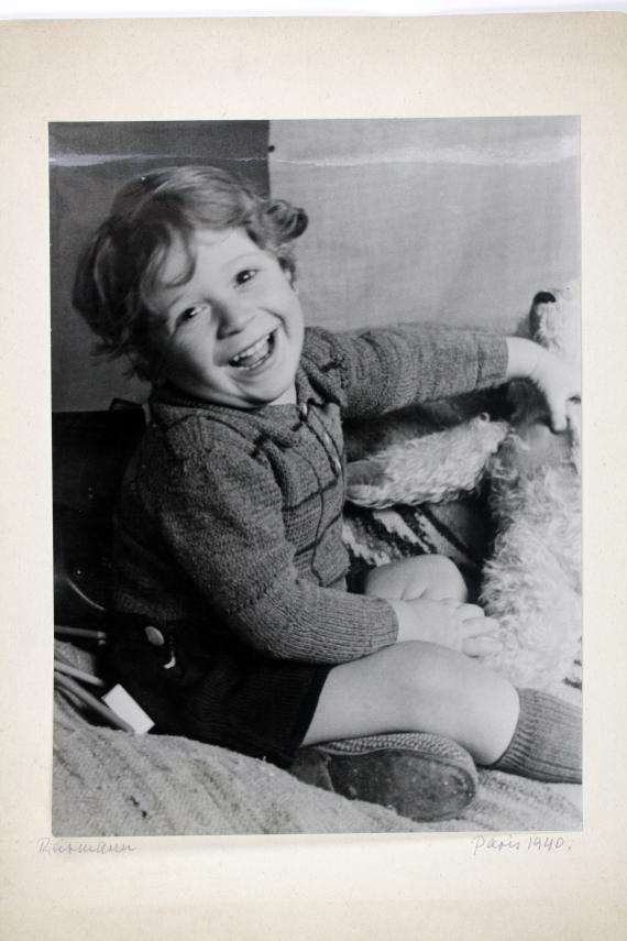 Schwarz-weiß-Fotografie, auf welchem ein Kleinkind auf dem Boden sitzend zu sehen ist. Vor ihm liegen Spielsachen auf dem Boden und er lächelt sehr fröhlich.