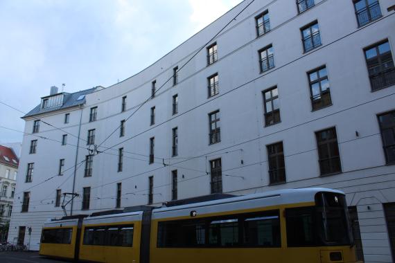 Breite und gerundete Fassade eines fünfstöckigen Hauses. Im Vordergrund eine Tram.
