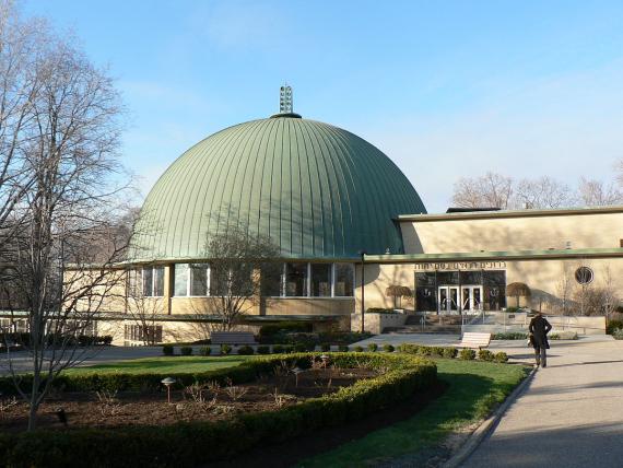 View over the park to a large dome of the synagogue, on the right a person, above the entrance Hebrew writing. In the background clear sky.